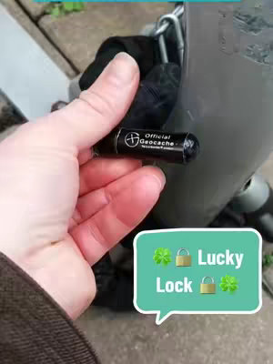 A person's hand holding a small, black geocache container next to a silver pole with a chain. The image also contains a speech bubble with icons and text.