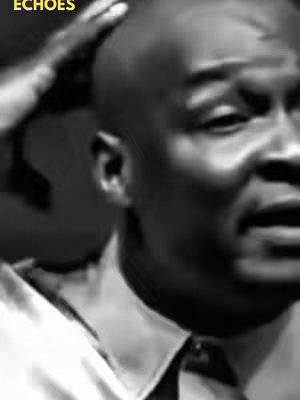 A black and white close-up portrait of a man, angled slightly towards the right. The man, who appears to be African, has a shaven head and is dressed in a collared shirt and tie. His expression is one of slight distress or concern. He is touching his head with his left hand. The background is a soft gradient of light and shadow.
