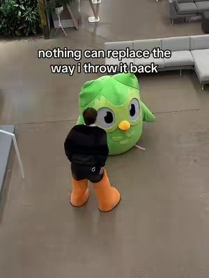 The image shows a person from behind, wearing a black outfit and orange boots, interacting with a large plush doll of Duo Lingo in a brightly lit setting. The text overlayed on top reads 'nothing can replace the way i throw it back'. The floor is gray and there is part of a couch visible in the background.