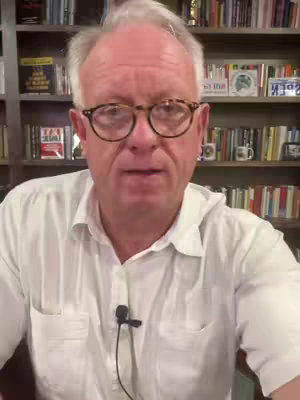 A middle-aged man with glasses and a white shirt is speaking directly to the camera. He is in front of a bookshelf filled with books.