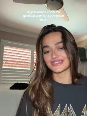 A woman with brown hair and a dark gray top is centered in a medium shot, with the text on the top of the view, inside a well-lit bedroom with a ceiling fan and a window, which has window blinds. The text reads, "so you wanna know the quickest way for a guy to run back"