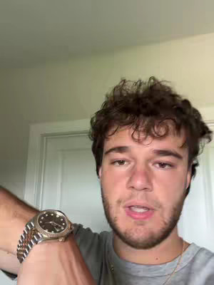 A man with short, curly brown hair is shown in a selfie-style shot. He is wearing a gray t-shirt and a gold necklace. A luxury watch is visible on his left wrist. The background includes a white door and a wall, suggesting he is indoors.