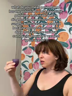 A woman with shoulder-length brown hair is looking to the left, mid-sentence, with a thoughtful expression. She's in a bathroom setting, with a colorful floral shower curtain in the background. She's holding a menstrual cup in her right hand, and wearing a black tank top. The text overlay on the image poses a provocative question.