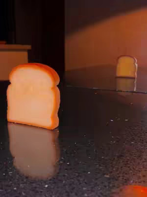 An eye-level shot features a slice of bread prominently in the foreground on a dark surface; another slice is further in the background creating depth, and reflecting in the table. The backdrop is dimly lit, suggesting an indoor setting. The surfaces have a dark, speckled appearance and the warm lighting emphasizes the bread. A slice in the back has a faint reflection.