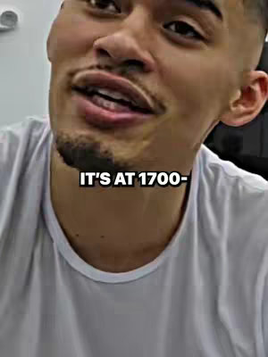 A medium close-up shot of a man with a light skin tone, wearing a white t-shirt. He is looking towards the left of the frame with a slight smile and his mouth open. A text overlay at the bottom reads, 'IT'S AT 1700-'. The lighting appears to be soft and diffused.