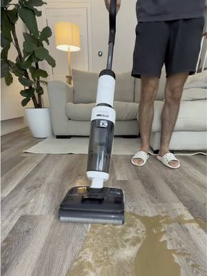 A person is shown using a wet and dry vacuum cleaner on a hardwood floor. The floor is partially covered with a brown liquid, simulating a spill. The vacuum cleaner is upright and held by a person. Behind the person there is a blurred background showing an apartment.