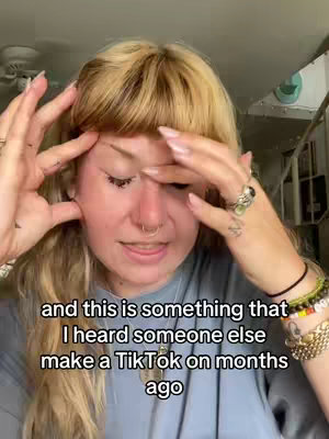 A selfie-style shot of a woman indoors, framed from the chest up. Her fair skin, with makeup subtly applied, contrasts with her natural blonde hair styled with bangs that frame her forehead and face. Her hands are positioned on either side of her head, with a finger touching her forehead in a gesture of thought. She wears a gray shirt, and her arms display various accessories, including rings, bracelets, and a watch, with tattoos on her wrists. The background is blurred but suggests an interior setting with what appears to be a ceiling fan and other items. The lighting is from above, casting gentle shadows on her face, enhancing the overall intimate and personal feel of the video.