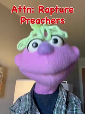A puppet with a purple head, bulbous nose, and large eyes is centered in the frame, wearing a plaid shirt. Overlay text in red arched format reads "Attn: Rapture Preachers". The background is an out-of-focus room.