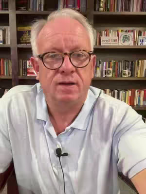 An older man in glasses is the central focus, positioned in front of a full bookshelf. The man wears a light blue collared shirt, and a microphone is clipped to it. The background is filled with a variety of books, creating a scholarly and informational atmosphere.