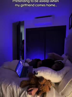 A bedroom scene with a man lying in bed, appearing to be asleep. A laptop is open on the bed, a teddy bear is beside him, and the room is illuminated by a blue light.