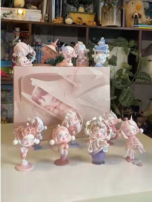 The image showcases an unboxing scene, focusing on various Skullpanda figurines displayed on a white surface. Several figurines are standing upright, with different poses and designs. A large box featuring an image of a figurine in a detailed presentation is behind them. The color palette consists of pink, off-white, and subtle variations of these colors, giving a delicate and aesthetic appeal. The background includes a wooden shelf filled with objects.