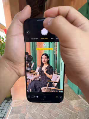 A person is filming their iPhone screen, displaying a photo of a woman seated at a cafe with green and orange color scheme. The person holding phone has large hands, and the phone's camera app is open with the capture button visible. Above the subject's head is a yellow text overlay. The background features a cafe setting.