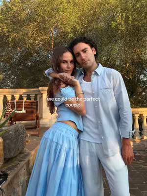 A couple is embracing each other while posing outside. The woman is wearing a sky-blue ensemble and has her arm affectionately around the man. The background is a soft focus of green foliage.