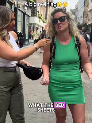 Two women are standing on a city street. One is holding a microphone and interviewing the other. The woman being interviewed is wearing sunglasses and a green dress. Text overlays say 'A big red flag' and 'WHAT THE BED SHEETS'.