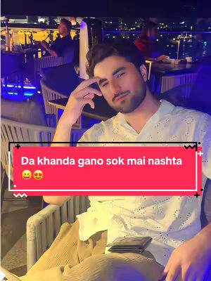 A man in a white shirt and beige pants sits in a chair at a restaurant or bar at night, with a red banner at the bottom containing text overlay. He is smiling with his hand near his face.