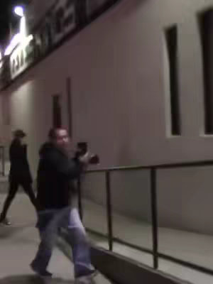 The image shows a man holding a phone in a street setting with another person walking in the background. The setting appears to be at night, as indicated by the lighting. The man in the foreground is wearing a black hoodie and jeans. The person in the background is less visible due to the lighting and distance.