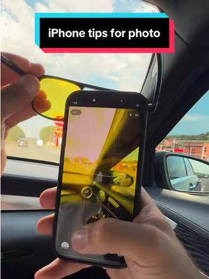 The image shows a person inside a car holding a smartphone, presumably filming. They are pointing a pair of yellow-tinted sunglasses towards the outside of the car, where a road and some buildings are visible. The phone screen reflects the car's interior and the sunglasses. The background shows a bright, sunny day.