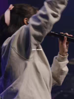 A person with dark hair and a blush-colored headband performs on a stage with a microphone in their hand. The person is wearing a white sweatshirt. Text overlay "ONE NAME" is displayed.