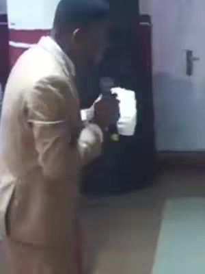 A man in a beige suit, slightly out of focus, holding a microphone. He is positioned to the left of the frame, speaking while facing slightly away from the camera. The background includes a dark form, possibly a speaker or equipment, and a white door with a handle.