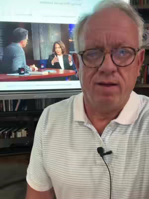 A man wearing glasses and a striped polo shirt stands in front of a TV screen showing Kamala Harris in an interview setting. The background includes a bookshelf.