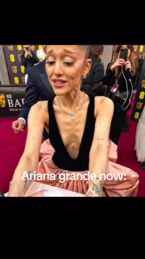 The image is a selfie-style shot featuring Ariana Grande. She is positioned slightly off-center, appearing to be in a public setting with a red carpet and individuals with cameras and microphones in the background. Ariana is wearing a black top combined with a pinkish/peach skirt, and a diamond necklace. Her expression shows visible emotion, with her mouth slightly opened and her eyes looking downward. The foreground is slightly out of focus. The text "Ariana grande now:" is placed at the bottom of the image.