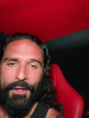 A man with long, dark hair and a beard sits in front of a red and black backdrop. Red racing style chair is visible and the text "BREAKING NEWS" is overlaid.