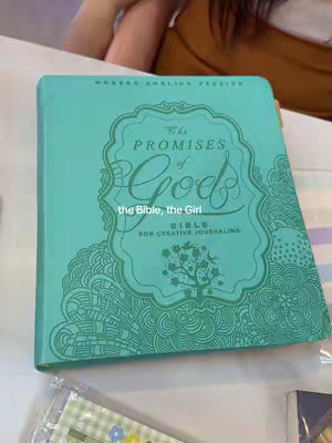 The cover image shows a Bible with a teal cover and decorative design, partially obscured by a book cover or table. The Bible's title, 'The Promises of God,' is prominently displayed. The overlay text says, "the Bible, the Girl."