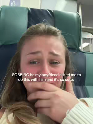 A close-up shot of a young woman crying, covering her mouth with her hand. She has blonde hair and is wearing a light-colored top. The background is a blurry image of a green and blue seat.