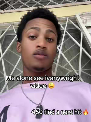 A close-up selfie of a young Black man with short, dark, curly hair. He is looking towards the camera with a neutral expression. The background includes a window with a white grid pattern. The man is wearing a light purple t-shirt with a chain necklace that has a black pendant.