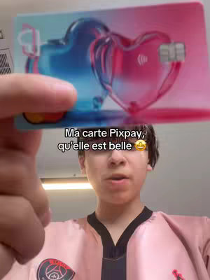 A selfie of a young man holding up a Pixpay card. The card has a heart-shaped design with pink and blue gradients. The man is wearing a pink shirt with a logo.