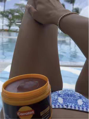 A person with tanned skin is applying tanning gel from a jar while sitting poolside. The jar is orange and contains brown gel. The person's leg is visible, with their hand holding the gel and applying it to their skin.