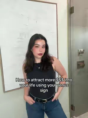 A woman in a black top and blue jeans stands in front of a white wall next to a glass shower. She is looking at the camera with a neutral expression and her hands are in her pockets. Text is overlaid on the image.