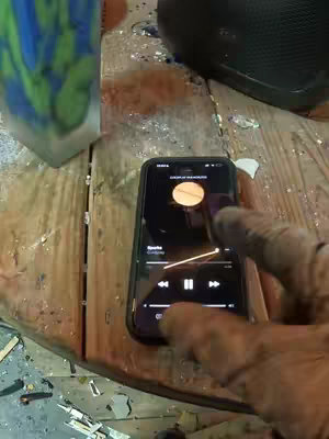 The image shows a smartphone displaying a music app interface, held by a hand wearing a glove. Beside the phone resting on a wood surface, is a partially visible, textured clear container. The background suggests a surface with debris, potentially a work area.