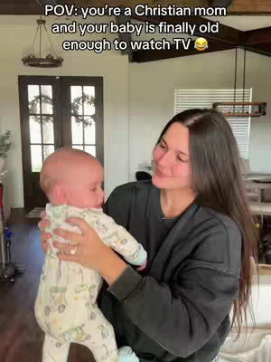 A woman with long dark hair holds a baby, looking down at the baby with a warm smile. The image is taken indoors, showcasing a comfortable home setting with a double door and ornate light fixture. The woman is wearing a black sweatshirt. The baby wears a light-colored onesie with a pattern.