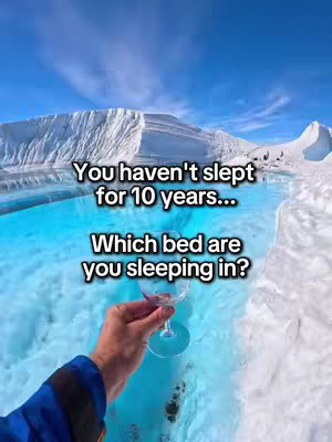 A first-person perspective shot showing a hand holding a stemmed glass with a scenic, snowy landscape in the background. The hand is positioned in front of a pool of blue water and a backdrop of snow-covered mountains and a bright blue sky.