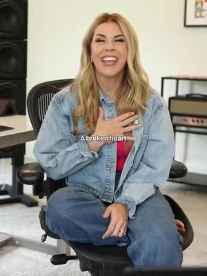 A blonde woman is sitting in an office chair, smiling at the camera and placing her right hand over her chest. The image has a light background and the woman is wearing a denim jacket.
