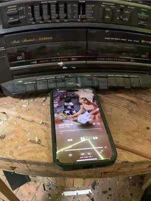 The cover photo shows a smartphone resting on a roughly textured wooden surface. The phone's screen displays a music app interface, with a photo of a woman wearing white on the screen. Above the phone, a black stereo system is in the background. The stereo's controls and speakers are visible. The overall composition is a cluttered but focused shot, with the phone as the focal point.