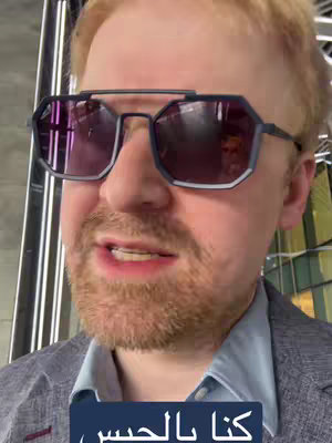 A close-up shot of a man wearing large, dark-rimmed sunglasses that reflect the surrounding environment. He has reddish hair and a beard. Below him is a blue text box with white text in Arabic script.