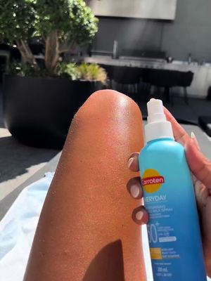 A close-up shot of a person's leg, likely a woman's, resting on a surface. They are holding a blue spray bottle of Carroten Everyday Moisturizing Milk Spray. The person has a light skin tone. The background is blurred.
