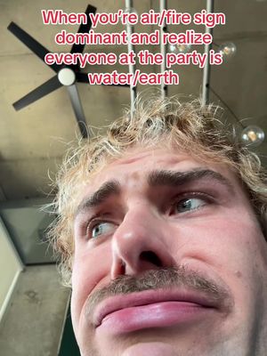 A selfie-style shot featuring a young man with curly blonde hair and a groomed mustache. He has a slight double chin from the upward perspective. Text overlays at the top read 'When you're air/fire sign dominant and realize everyone at the party is water/earth'. The background is a modern, slightly blurred interior with ceiling fixtures and lights.