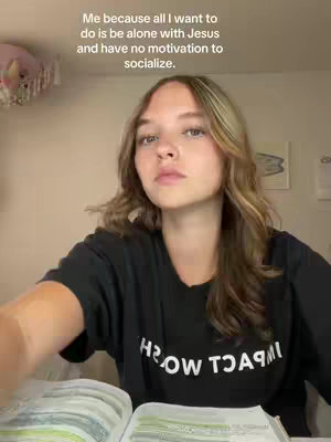 A young woman looks directly at the camera, sitting at a desk with a book open in front of her. She is wearing a black t-shirt, and text is overlaid at the top of the frame.