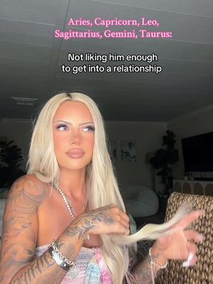 A woman with long, blonde hair is seated indoors, looking slightly to the side. She is wearing a low-cut top and a beaded necklace. Her arms are adorned with tattoos and bracelets. Text overlays are visible at the top and middle of the image. The background is softly lit, featuring hints of a living room setting with a couch and wall art.