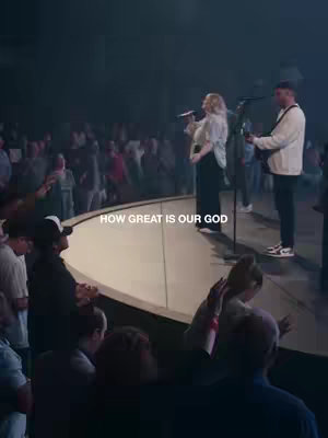 A live worship service featuring a male and female singer on a stage with a band and a large audience. The lighting is dim, creating a moody atmosphere. The text overlay at the bottom reads 'HOW GREAT IS OUR GOD'.
