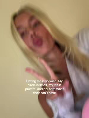 The image is a blurred selfie with a person, possibly a female, out of focus. The person seems to be making a peace sign gesture with their hand. Overlayed text is visible in the lower center area, beginning with the phrase 'Hating me is so valid.' The background is a light, solid color.