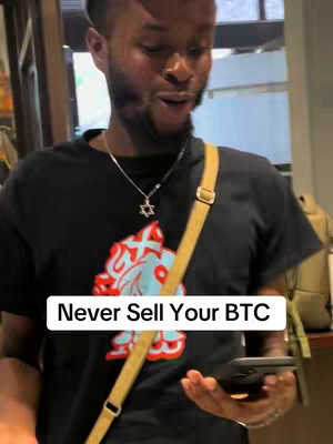 A man of African descent is the central figure, filmed from a medium-close distance. He's wearing a black t-shirt with a colorful design on the front and a gold shoulder bag diagonally across his body. A silver Star of David necklace hangs around his neck. The text overlay, on a white background, reads 'Never Sell Your BTC.' He is holding a black smartphone. The background remains slightly out of focus, likely an interior setting. The overall composition focuses on the man's upper body, highlighting his gesture and the text overlay.