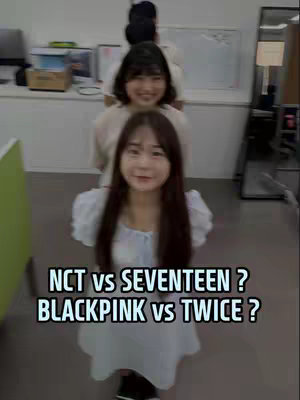 The image shows three young women standing in a line, viewed from a low angle, with the front woman being the closest to the camera. Above them, white text overlays read, \"NCT vs SEVENTEEN? BLACKPINK vs TWICE?\". The background appears to be an office setting, with a white board visible and some office equipment. The overall composition is straightforward, with the women taking center stage.