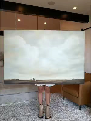 A person stands in front of a large rectangular painting, obscuring their torso and wearing a white dress and tan boots. The painting features a muted landscape with a person in the distance under a cloudy sky. The background includes a neutral-colored room with a rug, a brown leather chair, and a black beam along the ceiling.