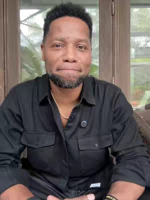 A man with a beard and a black shirt looks directly at the camera with a slightly concerned expression. Behind him, there's a blurred background suggesting an outdoor setting.