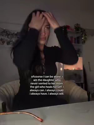 A young woman with long dark hair is in a dimly lit room, holding her hands up to her head. The woman is wearing a black long-sleeved top. Text is overlaid on the image.