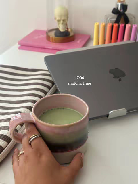 The image shows a close-up view of a hand holding a mug filled with a green matcha beverage. The mug is two-toned, with pink and green hues. The background includes a laptop, a decorative glass dome with a skull inside, books, and markers. The overall setting appears to be a study or workspace.